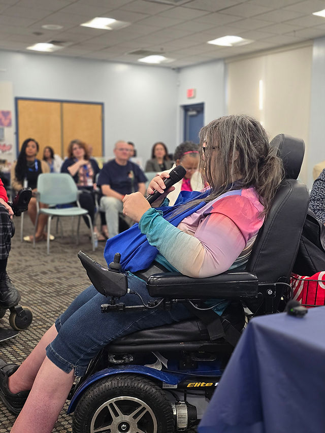 A white woman in a power wheelchair holds a microphone in her hand while speaking to the audience seated in the room. She has long, graying brown hair and is wearing a multi colored shirt with denim crop pants and black sneakers.