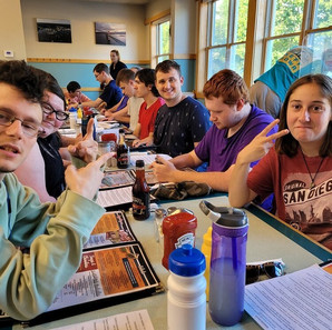 Several teens and young adults are seated at white formica tables in a restaurant. A few are facing towards the viewer and a young woman in the front right is making a peace sign gesture with her hand. The tables are scattered with menus, papers, condiment, and water bottles. There is a long row of plate glass windows in the background.