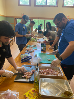 Trays of ingredients are laid out along a row of long wooden tables. Teenagers and adults are standing along the table cutting ingredients and prepping a meal.