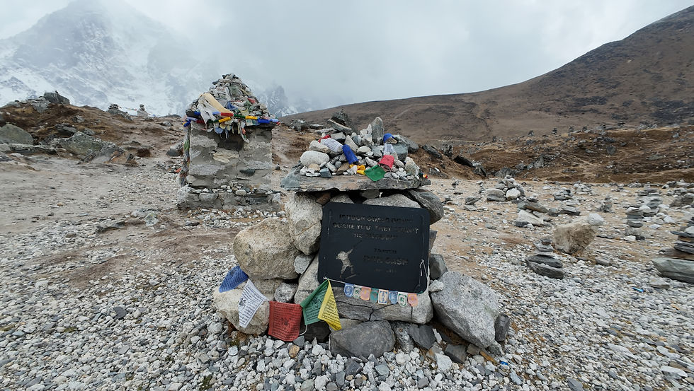 Rock memorial with colorful prayer flags in rocky mountain landscape. Plaque text faded. Snowy peaks in cloudy background convey solemn mood.