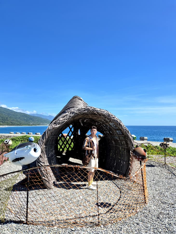 Charles stands inside woven sculpture by the sea under a clear blue sky. Shoreline and grassy hills in the background; colorful buoys nearby.