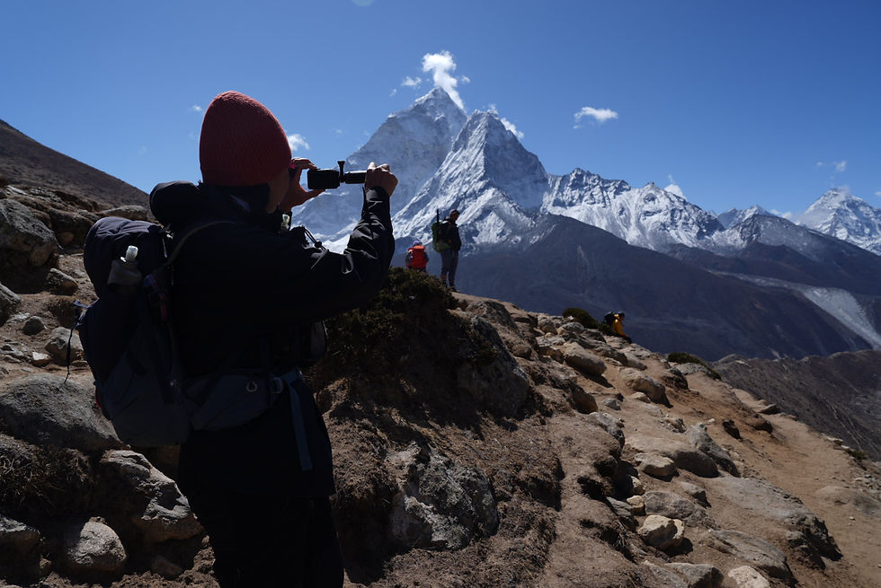 A person in a red hat takes a photo of snowy mountains. Two hikers in the distance, clear blue sky, rugged terrain creating an adventurous mood.