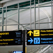Airport signs in Bahasa Melayu and English show directions for check-in, departure gates, arrival pick-up, and customer service.