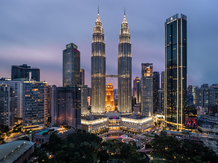 Kuala Lumpur skyline at dusk featuring the illuminated Petronas Twin Towers and surrounding skyscrapers, with a hazy purple sky.