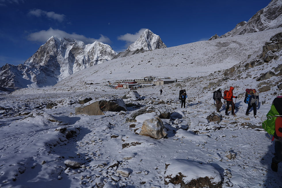 Hikers in colorful gear trek through a snowy mountain landscape with snowy peaks and a small settlement in the background under a clear blue sky.