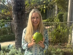 woman with blonde hair stands outside surrounded by plants smiling holding coconut