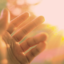 A hand reaches toward a bright, golden sun, with rays illuminating a blurred, green background. Warm, serene mood.