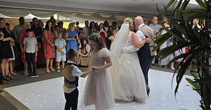 Bride and groom dance at wedding; guests watch on a white dance floor.