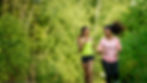 Two women smiling while jogging together on a wooded trail.