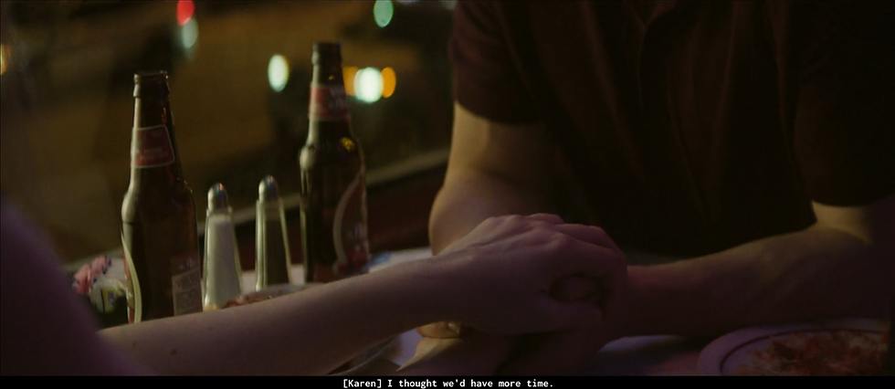 Two people hold hands across a table in a dimly lit restaurant with beer bottles. Subtitles read: [Karen] I thought we'd have more time.