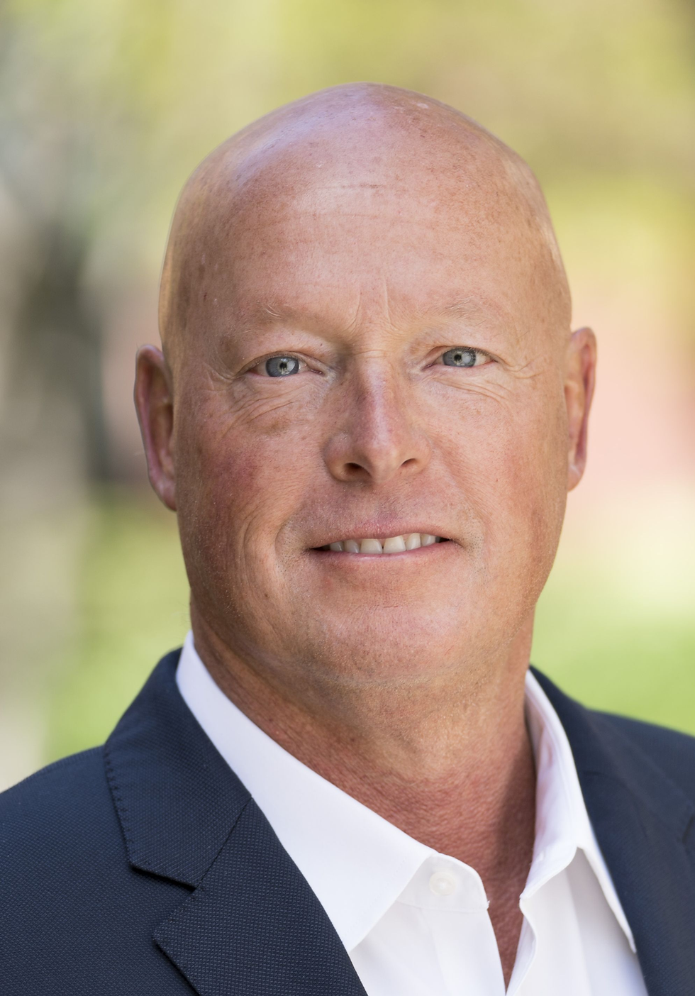 Close-up portrait of a bald man in a navy suit and white shirt, slight smile, against a soft blurred outdoor background.