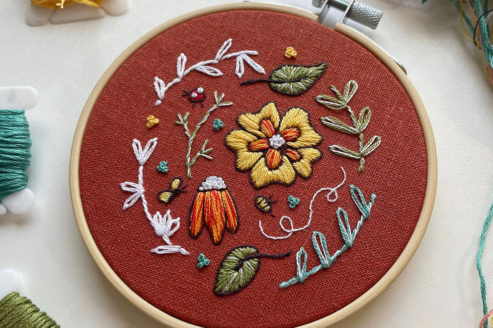 Photo of an embroidery hoop with a floral sampler stitched in yellow, red, white, blue, and green onto dark orange fabric.