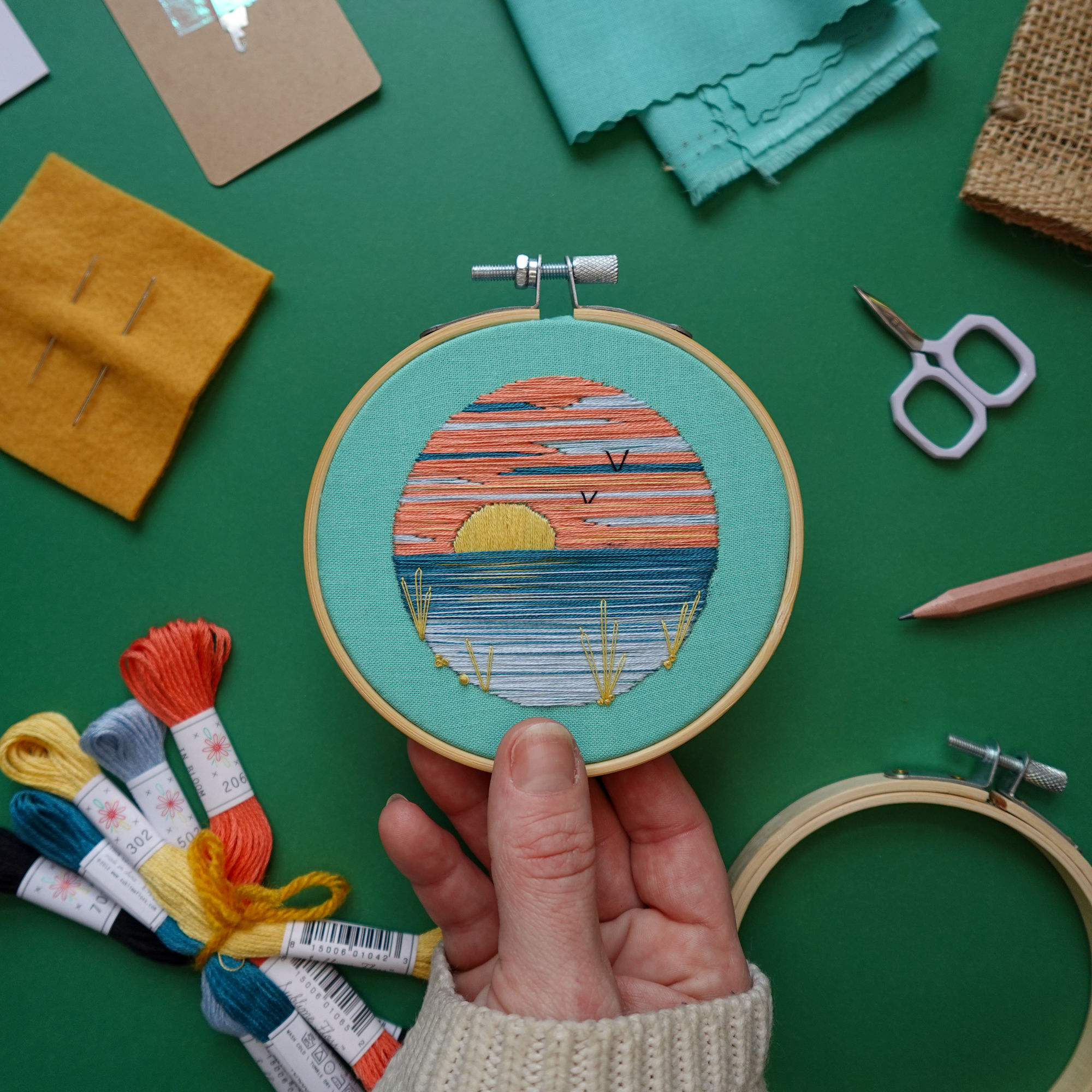 Embroidery project featuring a colorful sunset over water and birds in the background, held over supplies.