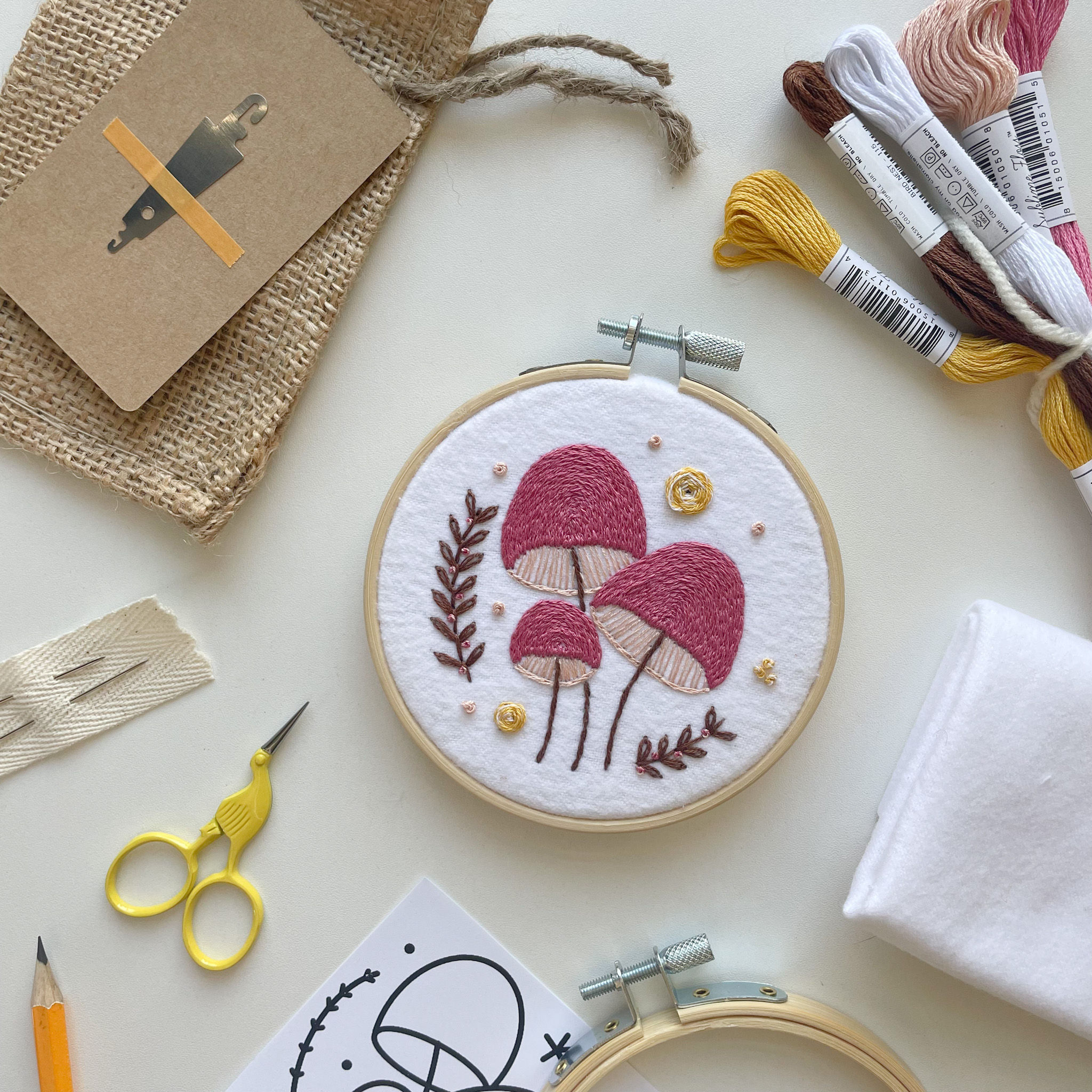 Pink mushroom embroidery project surrounded by various embroidery supplies on a white surface.
