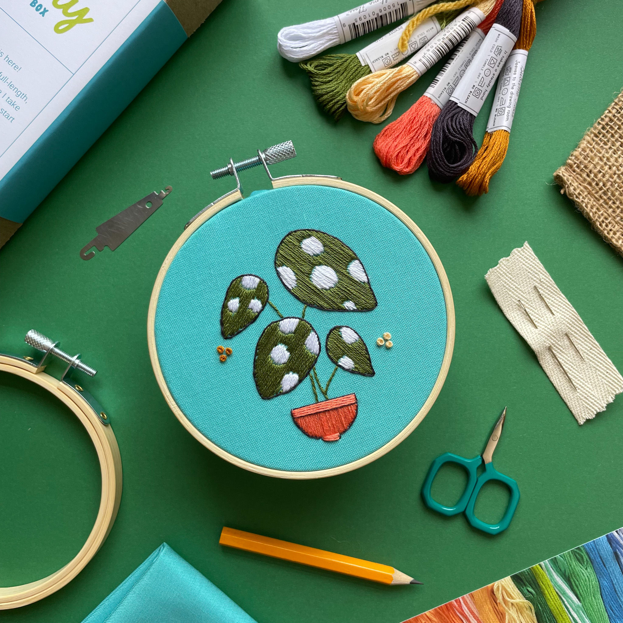 Embroidery hoop with a green and white spotted begonia plant stitched onto a bright blue cotton fabric.