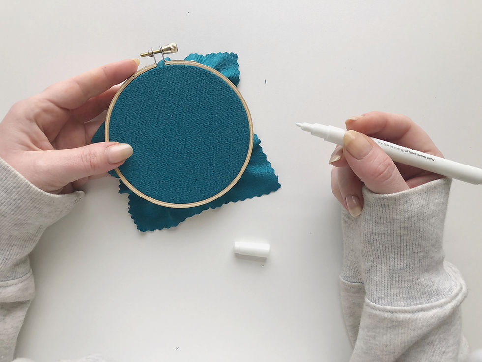Hope holds an embroidery hoop with dark blue fabric in one hand, and a white pen in the other.