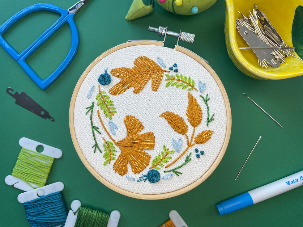 Embroidery hoop with three leaves surrounded by vines and flowers stitched onto off white fabric. Hoop is surrounded by various embroidery supplies.