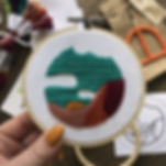 An embroidery hoop with a landscape stitched in maroon, deep orange, pink, and blue on white fabric.