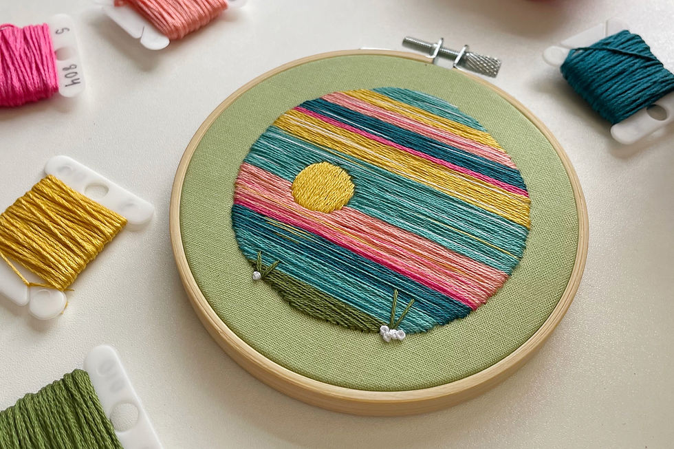 A photo of an embroidery hoop with a landscape stitched in blues, greens, yellows, and pinks onto light green fabric.