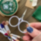 Thumbnail: Photo of an embroidery kit for a hoop featuring white snowflakes stitched onto green fabric, surrounded by winter foliage.