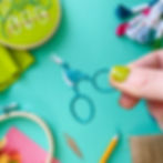 Embroidery scissors shaped like storks in a green color, held over a blue surface with scattered embroidery kit supplies.
