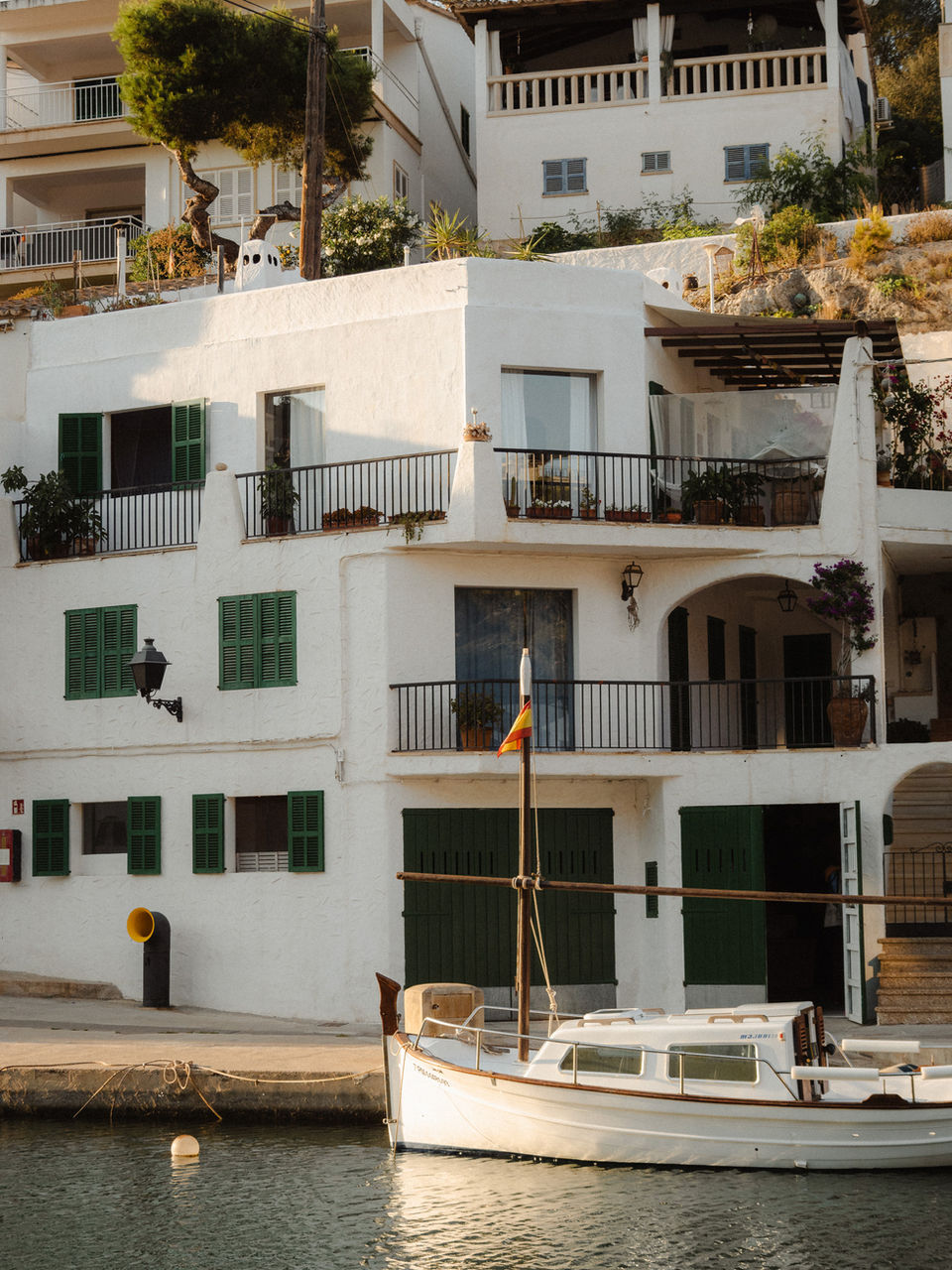 A white boat is slightly illuminated by the setting sun. it is tranquilly anchored in a tiny harbor in front of a mediterranean white house with many green doors and window-blinds. This is an image from the detailpage “Mallorca” on the website Fabian Wiktor Photography.