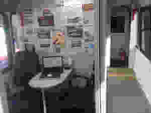 Parte interna do ônibus Lilás, com uma mesa pequena com um computador ao centro e duas cadeiras