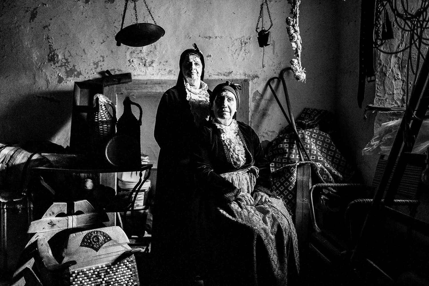 Two elderly women inside an abandoned warehouse at Portaria, wearing the traditional costumes of Pelion, Thessaly, Greece. Black and white photography by George Tatakis