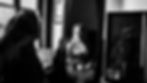 Black and white close-up portrait of a young woman in Soufli traditional attire gazing into an antique mirror. Her face is illuminated as she meets the camera, with her embroidered headscarf and coin necklace visible, conveying a reflection on cultural identity.