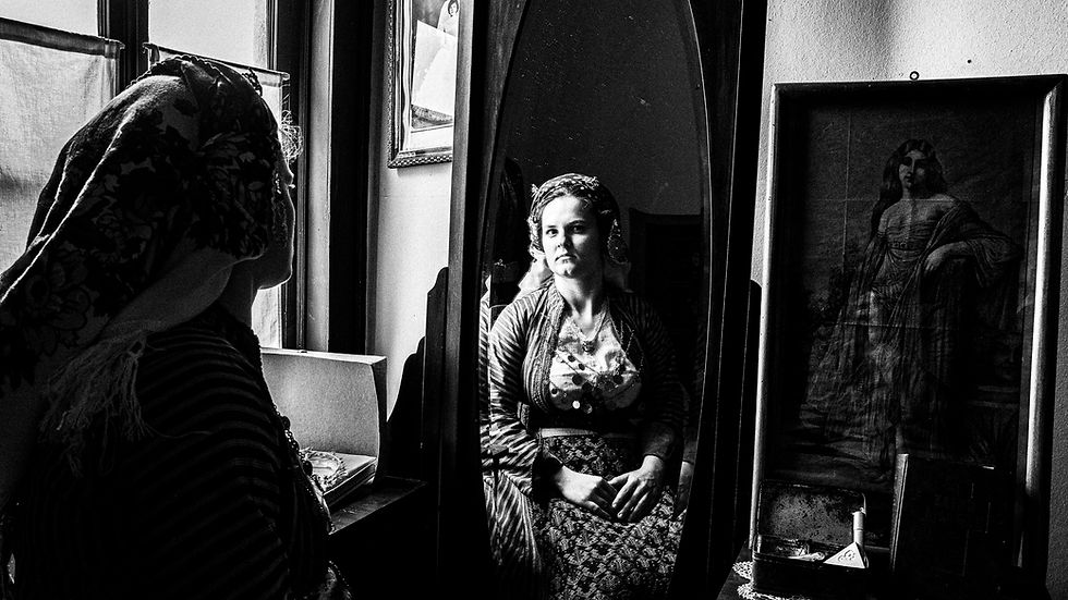 Black and white close-up portrait of a young woman in Soufli traditional attire gazing into an antique mirror. Her face is illuminated as she meets the camera, with her embroidered headscarf and coin necklace visible, conveying a reflection on cultural identity.