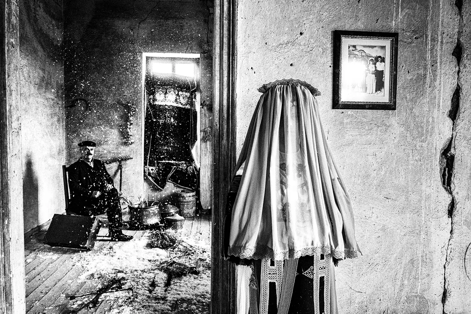 A bride wearing the Karagouna bridal dress with her husband in the background inside an old mansion in Glinos, Trikala, Thessaly, Greece. Black and white photography by George Tatakis