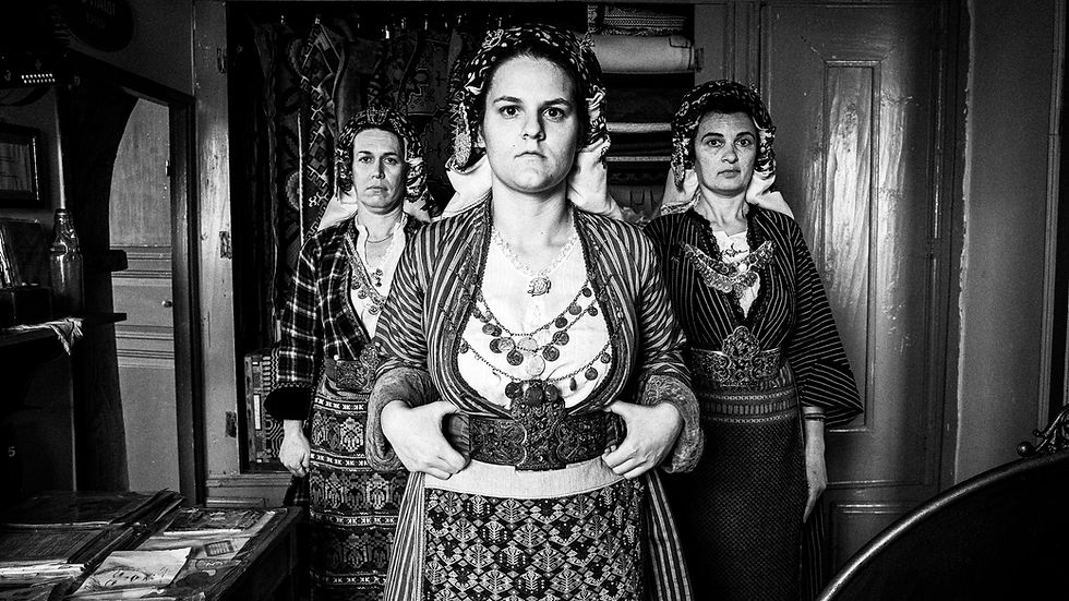 Black and white photograph of three Soufli women standing shoulder to shoulder in full traditional costume against a doorway backdrop. They face the camera with proud expressions, showcasing detailed textiles, belts, and jewelry of Thracian folk dress, representing the guardians of local tradition.