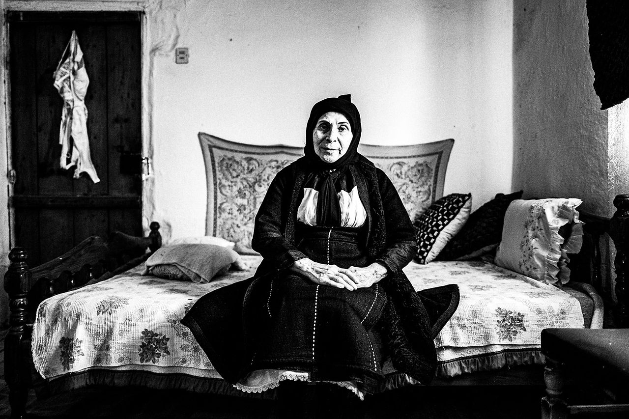 An elderly woman wearing the local folklore costume, sitting on a bed at a traditional local home. Meliki, Imathia, Macedonia, Greece. Black and white photograph by George Tatakis