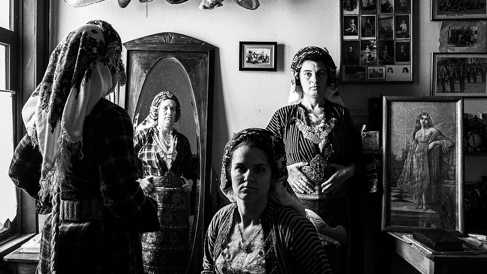 Black and white photo of three Soufli women in traditional dress inside a historic room. One woman’s reflection appears in a tall mirror as another stands beside it; old photographs and artwork decorate the walls, highlighting the reflection of past and present.