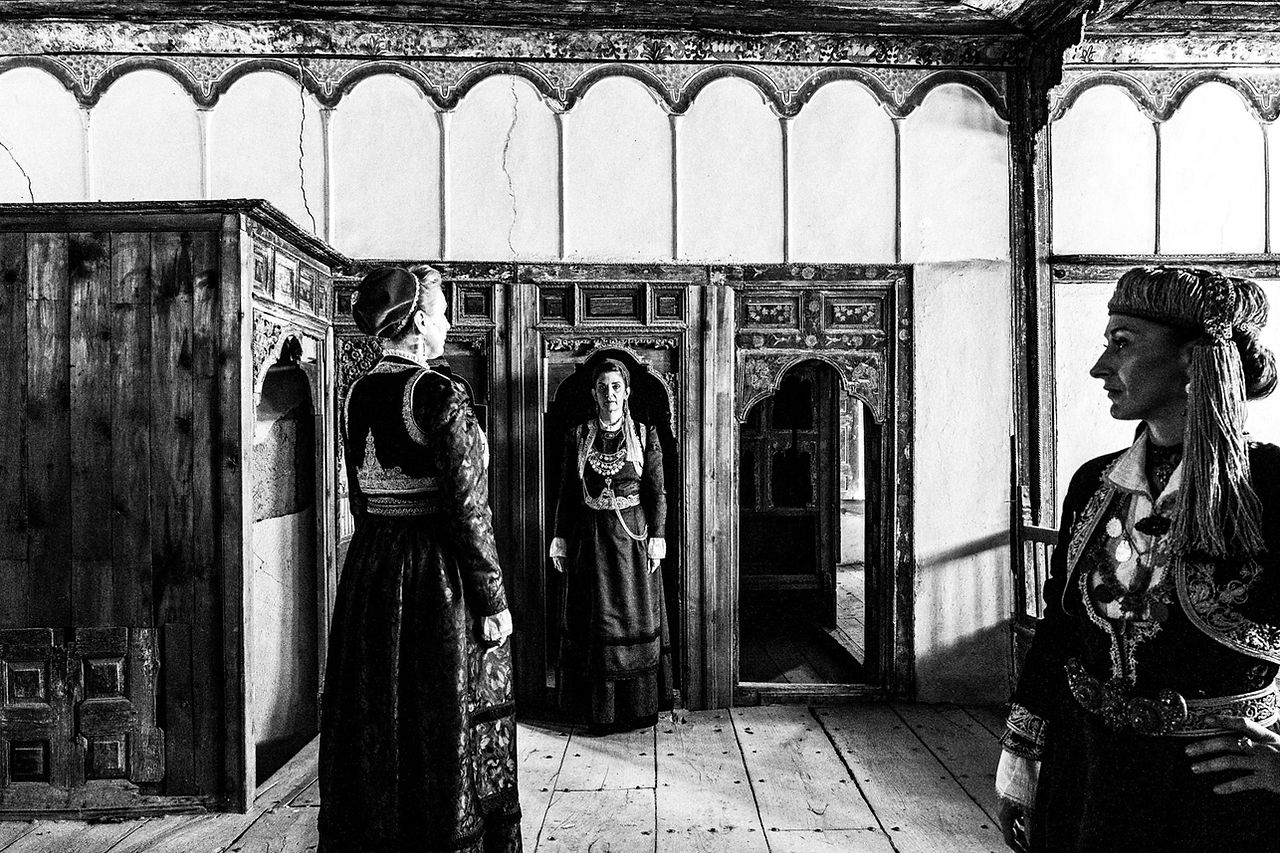 Three women standing inside a Mansion in Siatista, wearing the urban dresses of Kozani, Macedonia, Greece. Black and white photograph by George Tatakis
