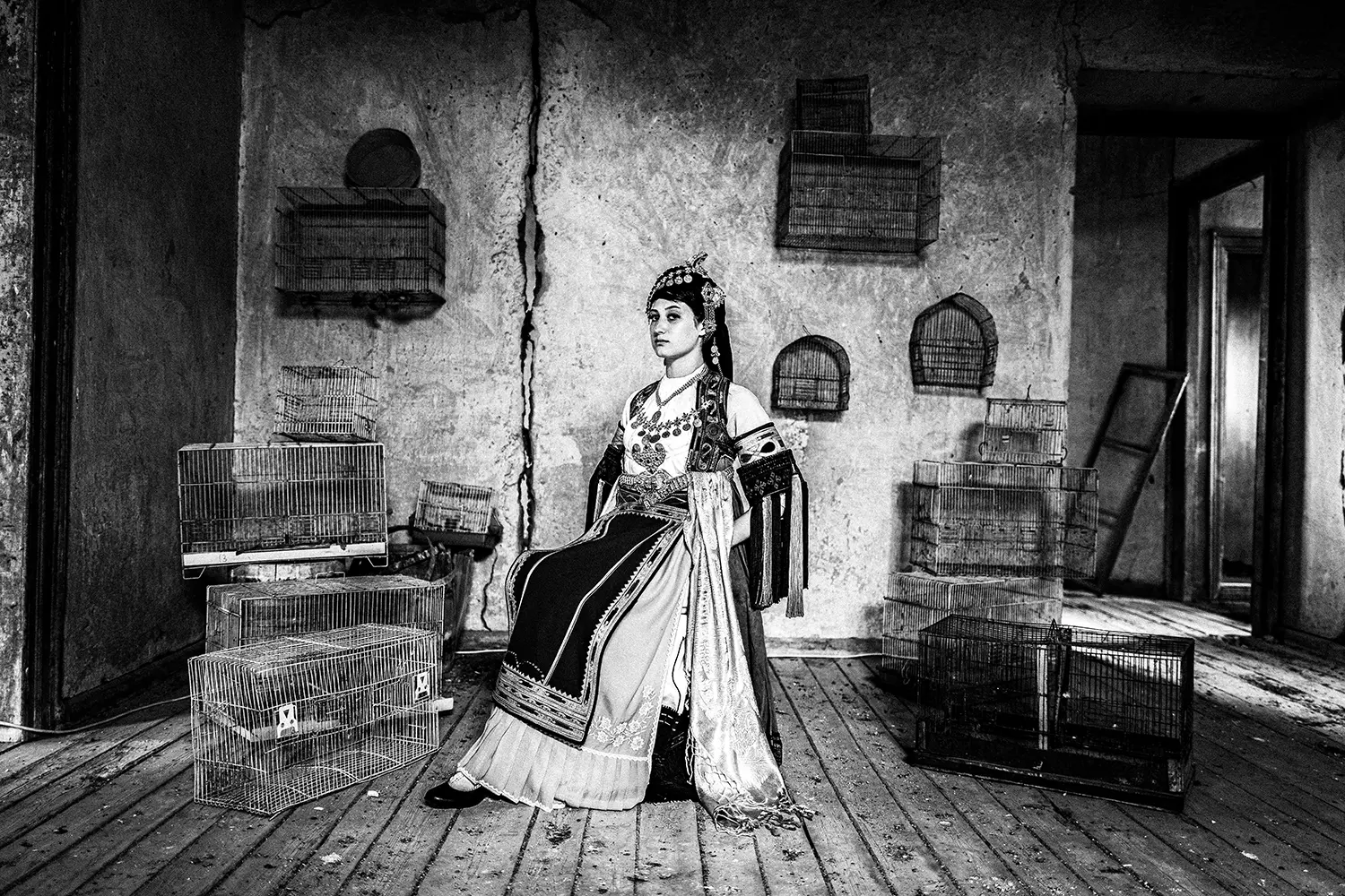 A woman inside a room full of bird cages, at a mansion in Glinos, wearing the traditional Karagouna dress. Trikala, Thessaly, Greece. Black and white photography by George Tatakis