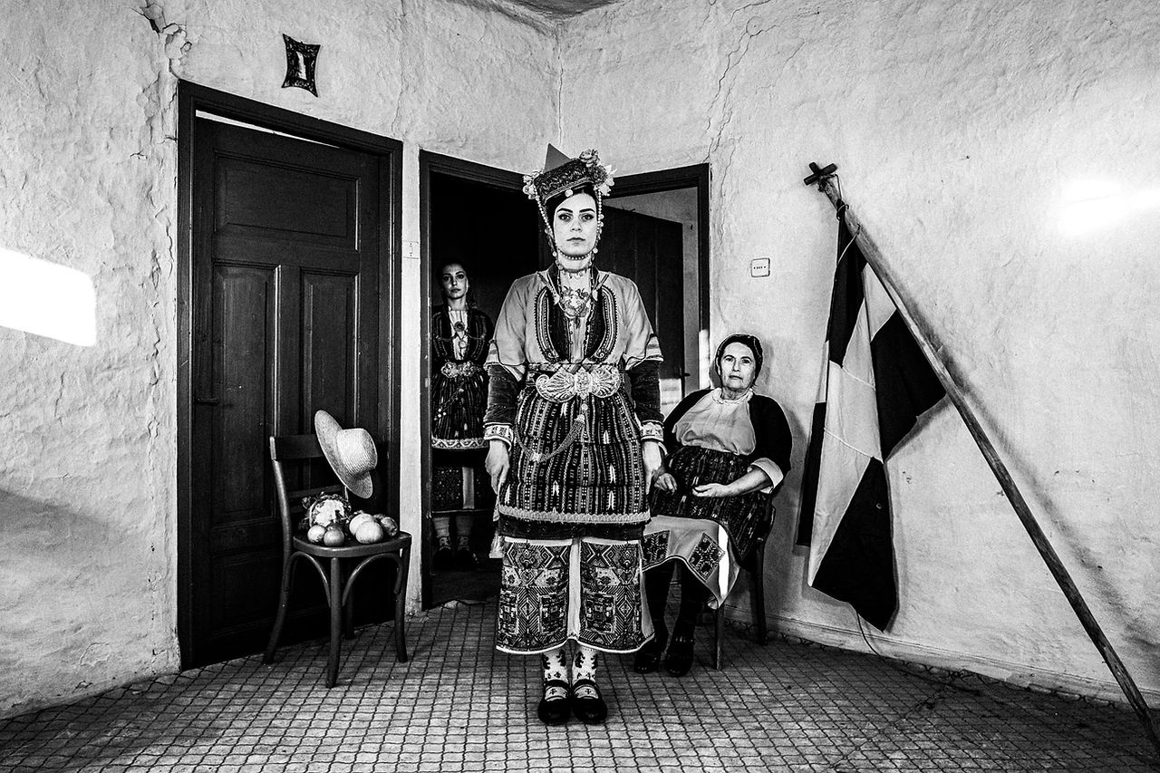 Three women at a local traditional home, in their traditional clothes, next to the Greek flag. Aggelochori, Naoussa, Macedonia, Greece. Black and white photograph by George Tatakis