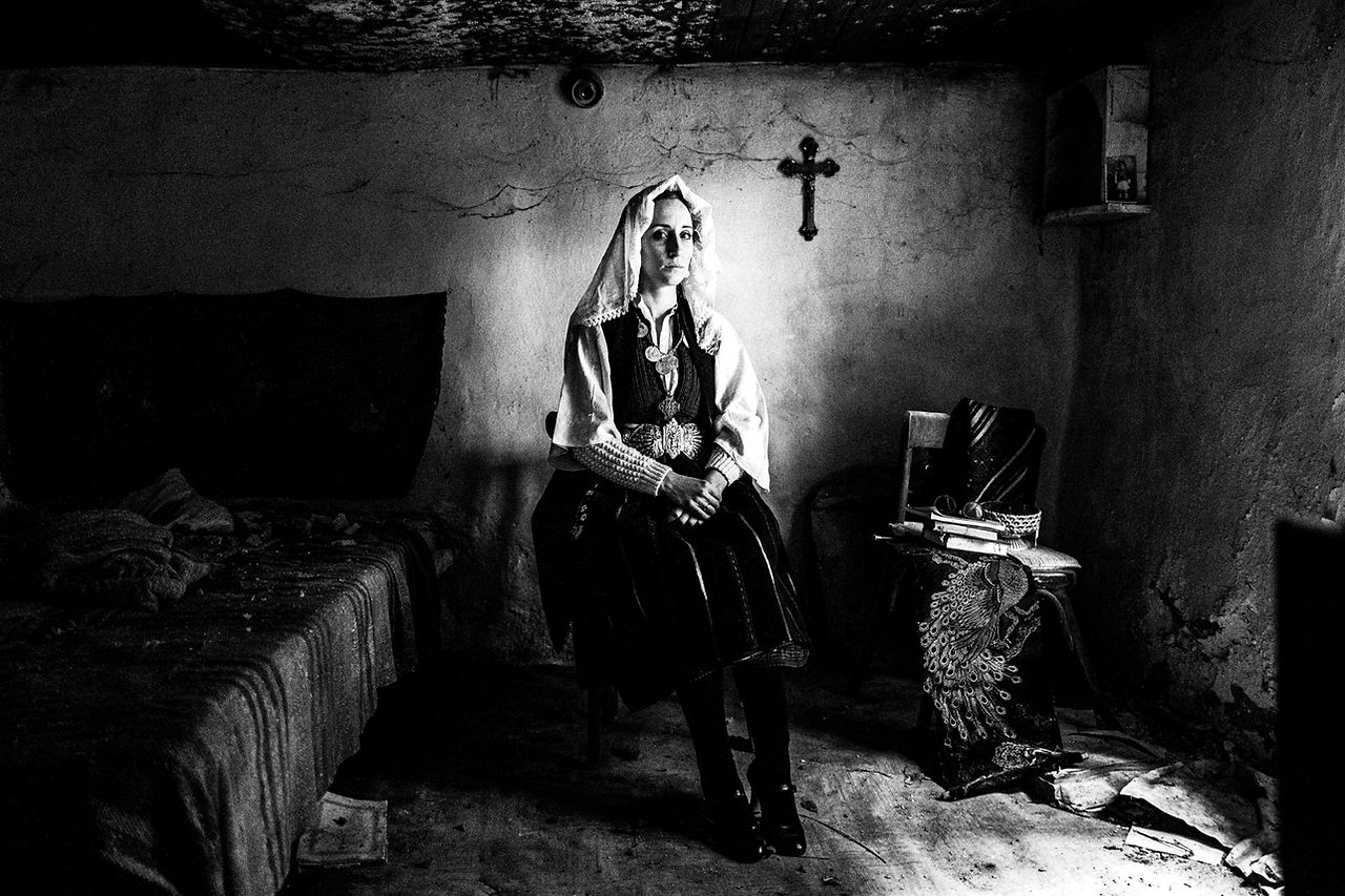 Sitting portrait of a woman in her traditional bridal dress, inside a local abandoned house. Ermakia, Ptolemaida, Macedonia, Greece. Black and white photograph by George Tatakis