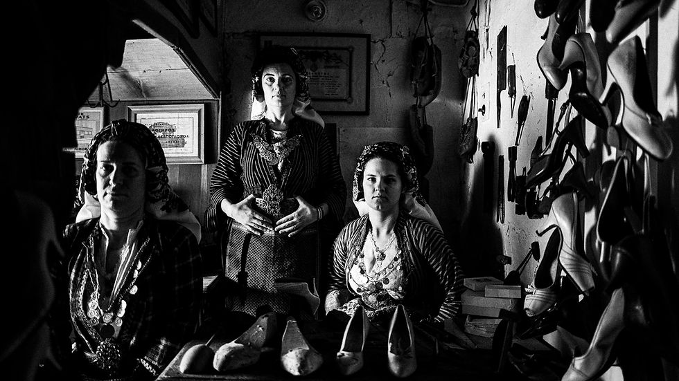 Black and white photo of three women in traditional plaid costumes in an old cobbler’s workshop. Vintage shoe forms, tools, and a Singer sewing machine surround them, illuminated by window light, capturing a timeless craft environment.