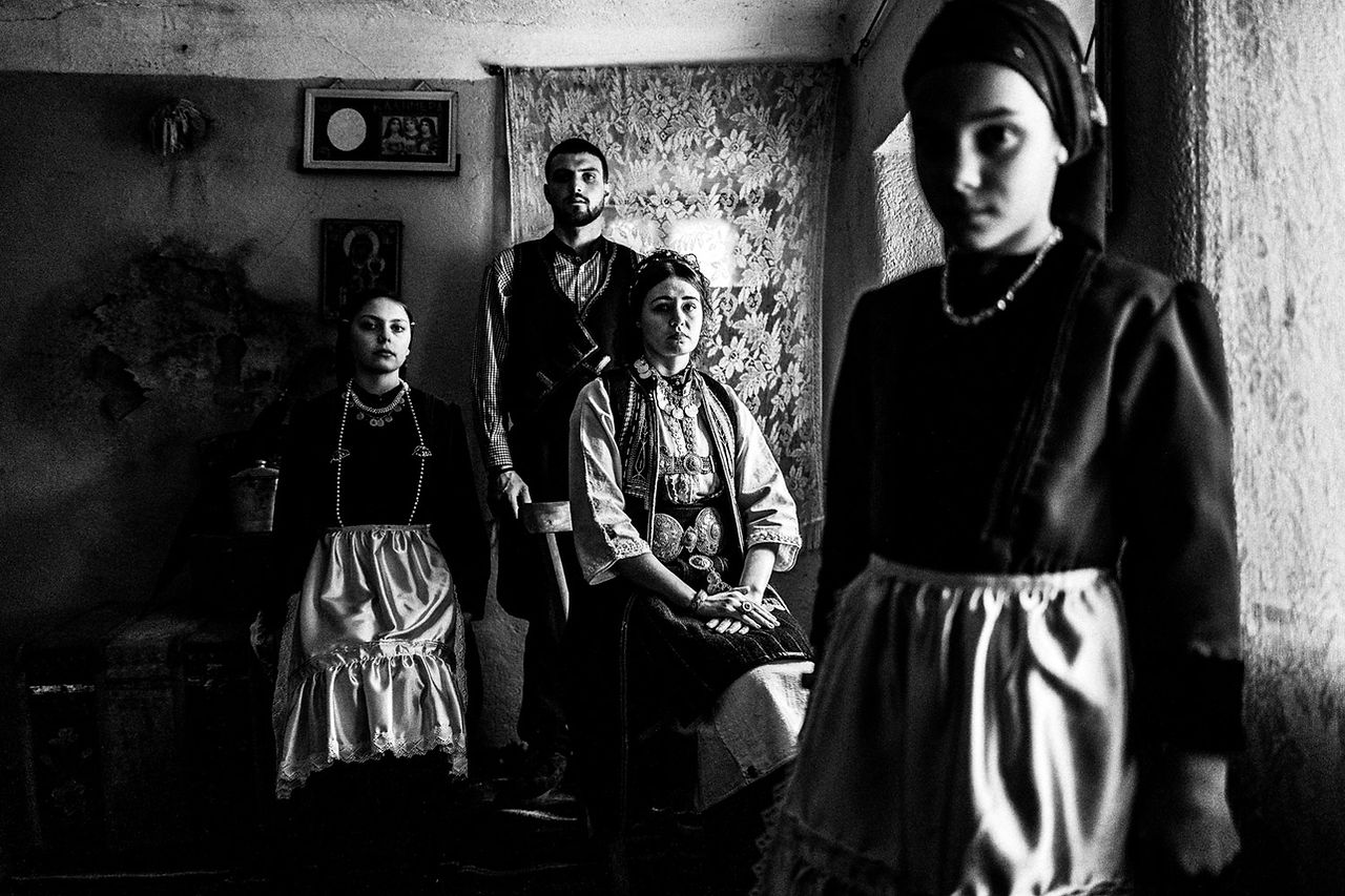 Four persons inside a local home, wearing the local attire. Polypotamos, Florina, Macedonia, Greece. Black and white photograph by George Tatakis