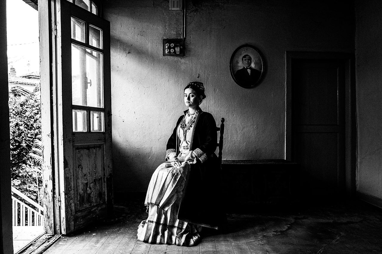 Sitting portrait of a woman inside a local abandoned house, wearing the traditional local attire. Veria, Imathia, Macedonia, Greece. Black and white photograph by George Tatakis