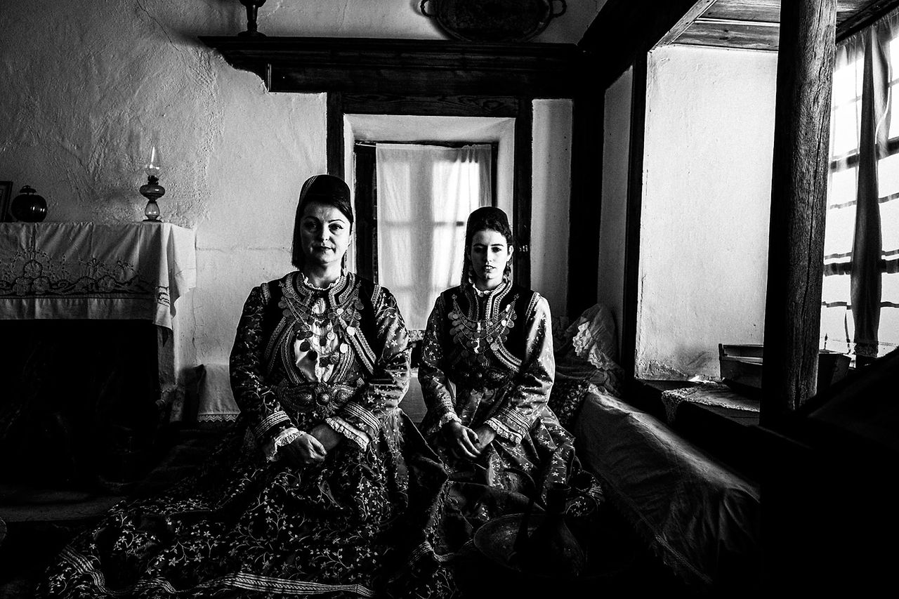 Two women inside the bedroom of a local Mansion, wearing the local folklore dresses. Kastoria, Macedonia, Greece. Black and white photograph by George Tatakis