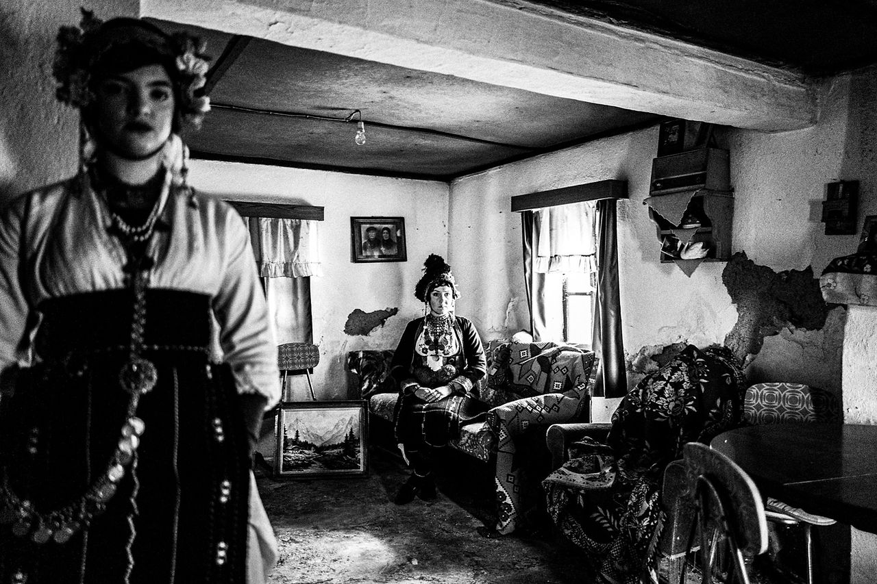Two women, wearing the traditional attire, posing inside a local traditional house. Meliki, Imathia, Macedonia, Greece. Black and white photograph by George Tatakis