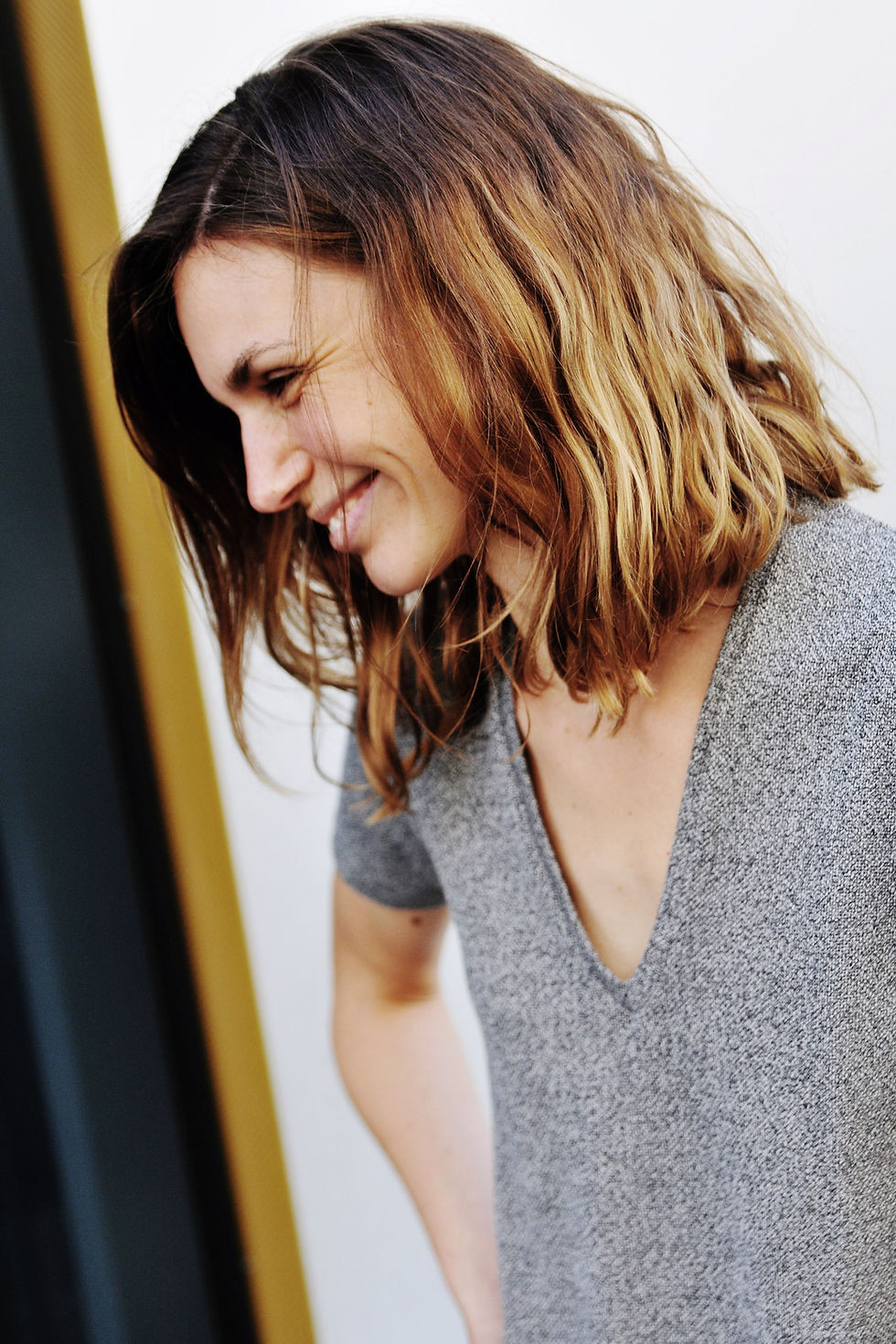 Auf dem Bild ist eine weiblich gelesene Person zu sehen, die seitlich nach unten guckt und lächelt. Sie hat lachende Augen, trägt ein graues T-Shirt mit V-Ausschnitt. Der Hintergund ist hell.