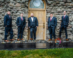 Groom and groomsman posed against stone wall barn setting.