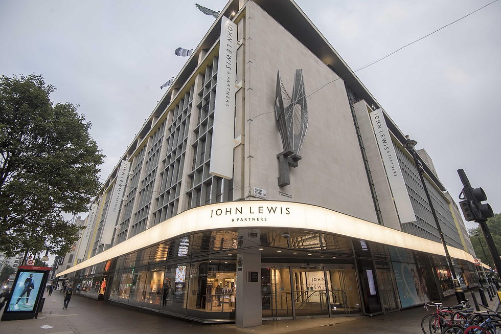Modern building facade of John Lewis & Partners with visible signage. Overcast sky, bicycles, and a digital ad panel on a busy street corner.