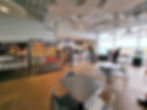 Silverstone museum cafe with tables and chairs, people ordering at a counter, shelves of drinks and snacks labeled "Grab & Go." Large windows, bright lighting.