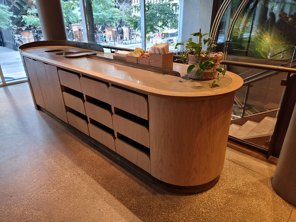 Wooden counter with slots, a plant in a woven basket, napkins, and utensils on top. Glass windows show a city street and trees outside.