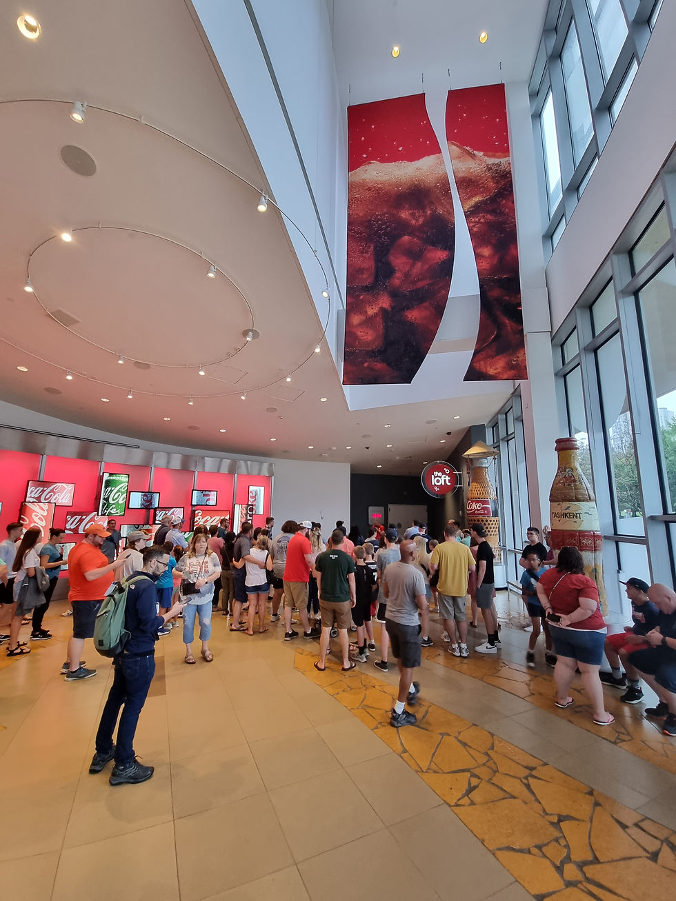 People in a spacious room with large Coke banners, standing in line. Bright lighting, red and white decor, and an excited atmosphere.
