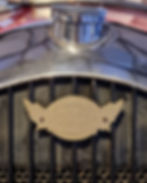 Close-up of a vintage car grille featuring a brass "Morgan" emblem with wings. Reflective silver cap above; background shows a red blur.